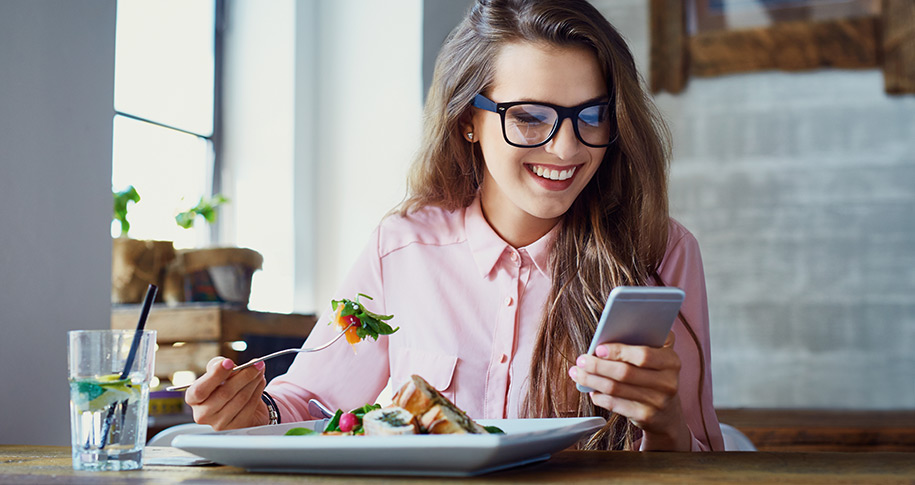 Chica con gafas comiendo comida saludable mientras mira su móvil.