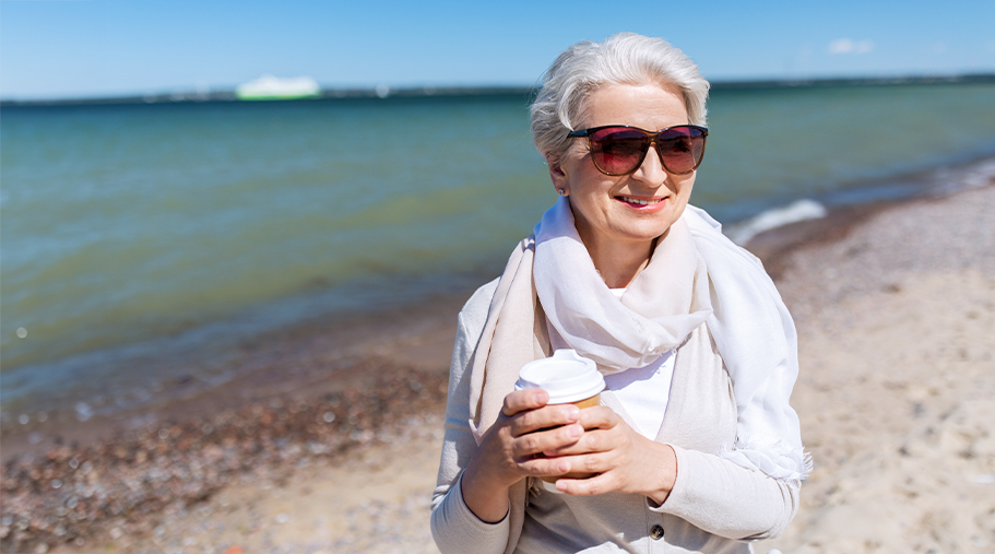 Mujer mayor paseando por la playa con gafas de sol y un café en las manos